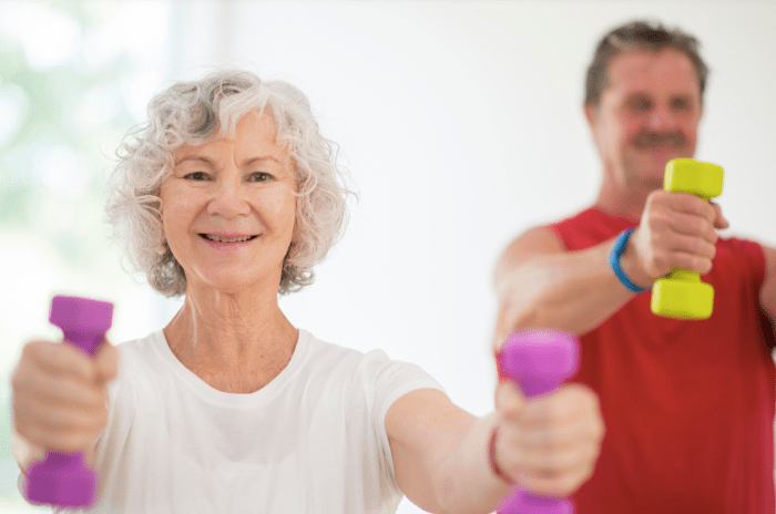 An older lady in a t-shirt is lifting small dumbells, with an older gent behind her. 